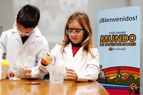 Niña y niño realizando experimento frente al banner de Mundo de Investigadores.