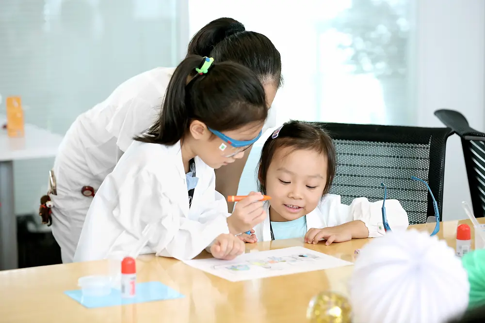 Dos niños y una mujer con bata de investigador colorean un cuadro en una mesa.