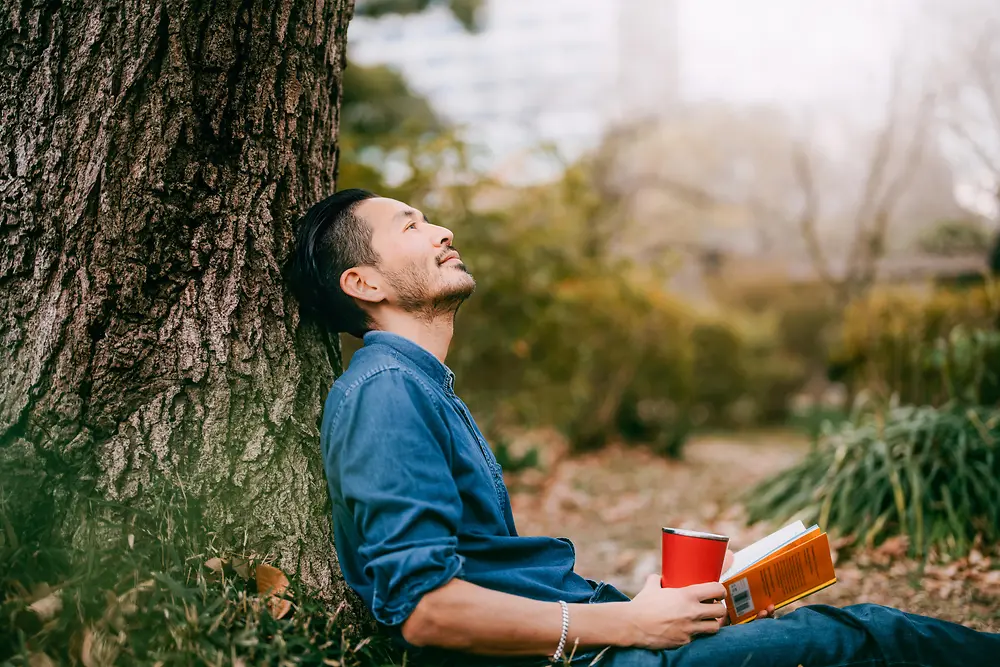 Un hombre con un libro recargado en un árbol.