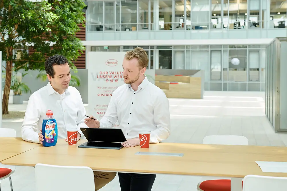 Dos hombres de pie a lado de una mesa con una botella de Persil y una computadora portátil.