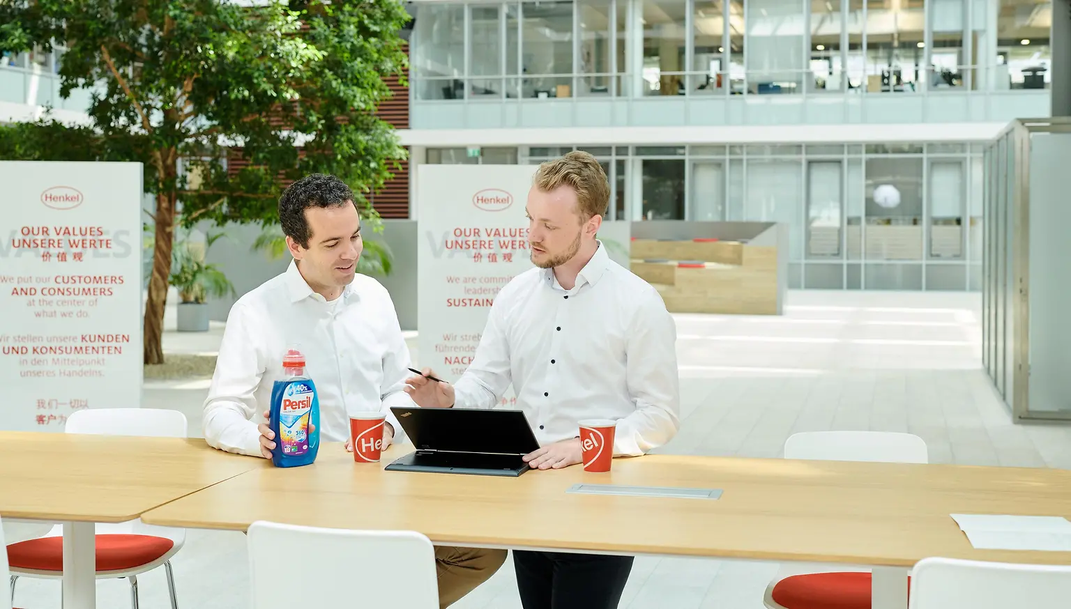 Dos hombres de pie a lado de una mesa con una botella de Persil y una computadora portátil.